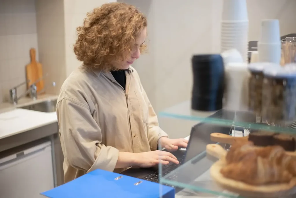Café owner working on a laptop at the counter, reviewing costs beside the pastry display